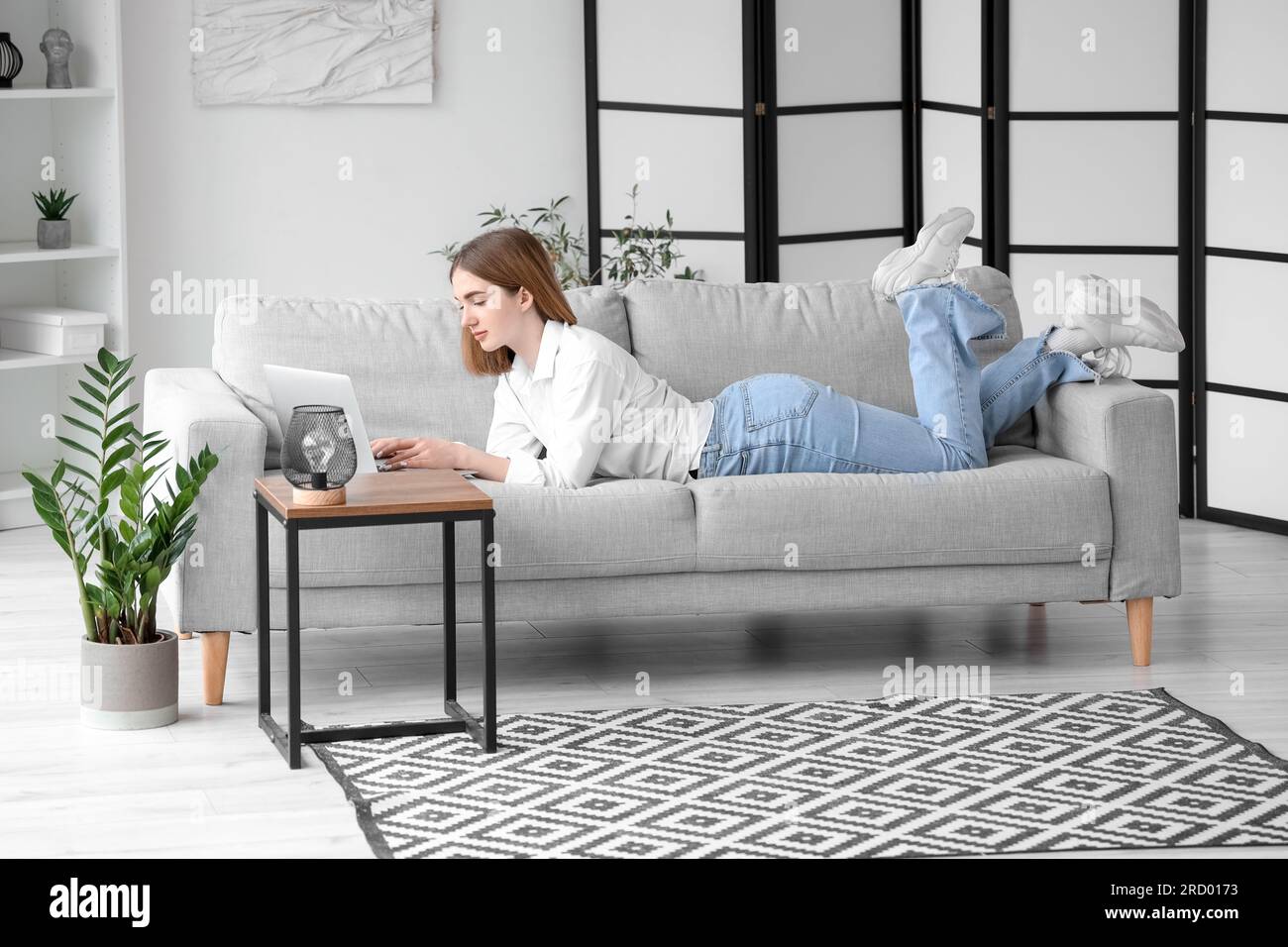 Pretty young woman lying on grey sofa and using modern laptop in light ...
