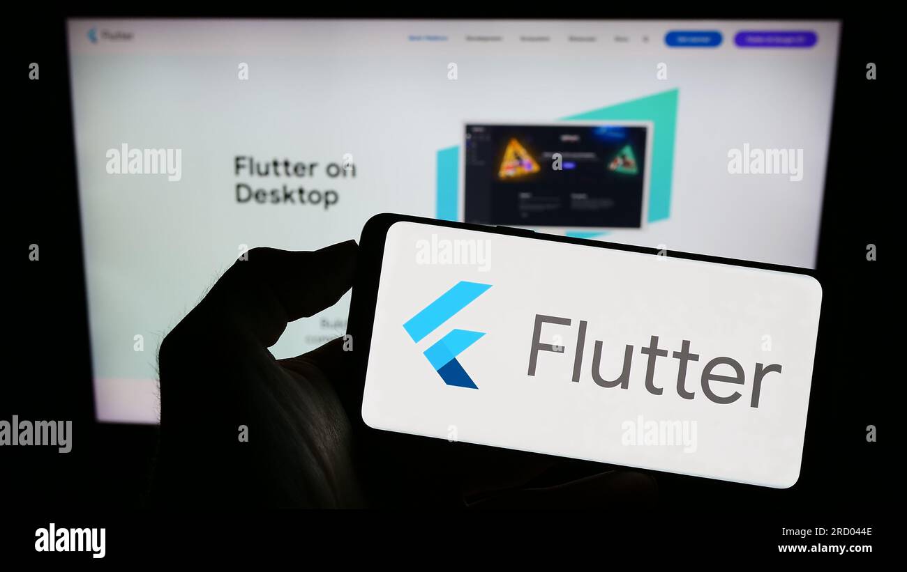 Person holding cellphone with logo of UI software development kit Flutter (Google) on screen in front of webpage. Focus on phone display. Stock Photo