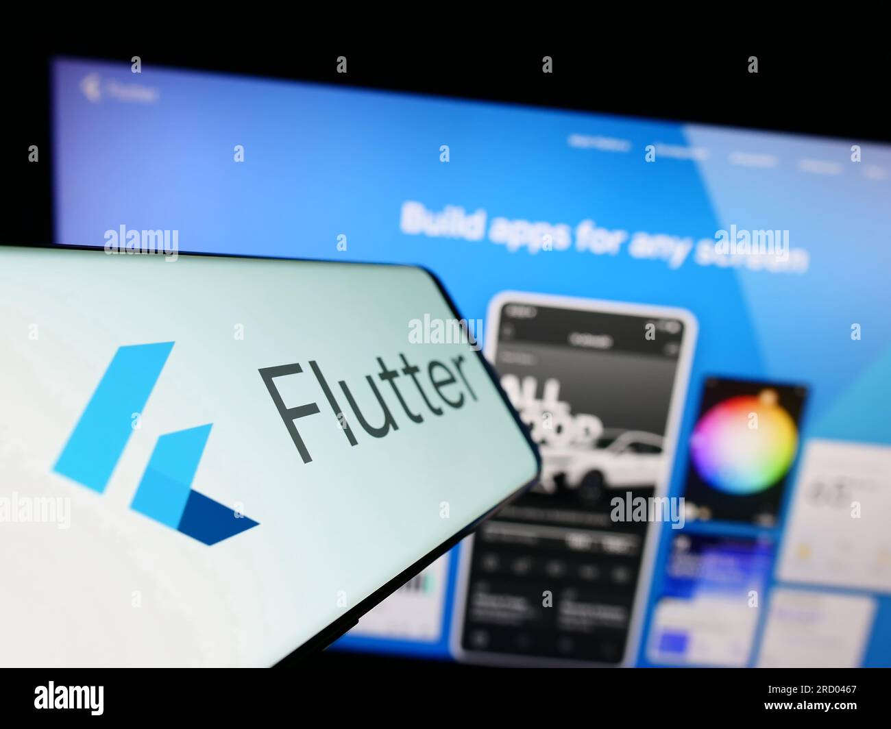 Mobile phone with logo of UI software development kit Flutter (Google) on screen in front of website. Focus on center-left of phone display. Stock Photo