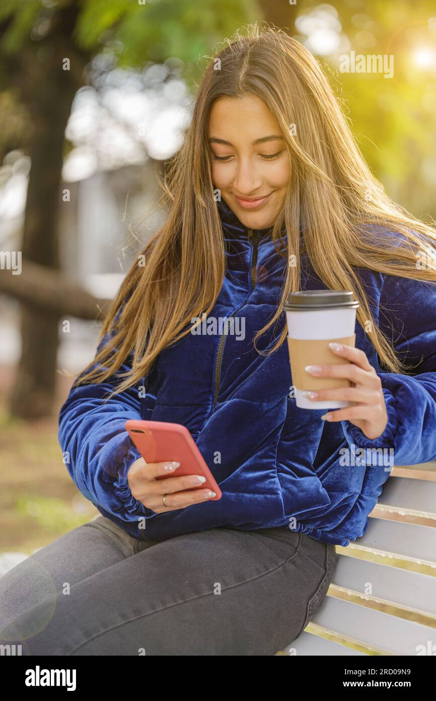 Latin girl sitting on a bench in a public park drinking coffee and using her mobile phone. Stock Photo