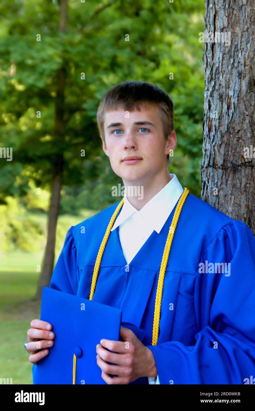 Young man stands wearing his blue graduation gown and holding his hat