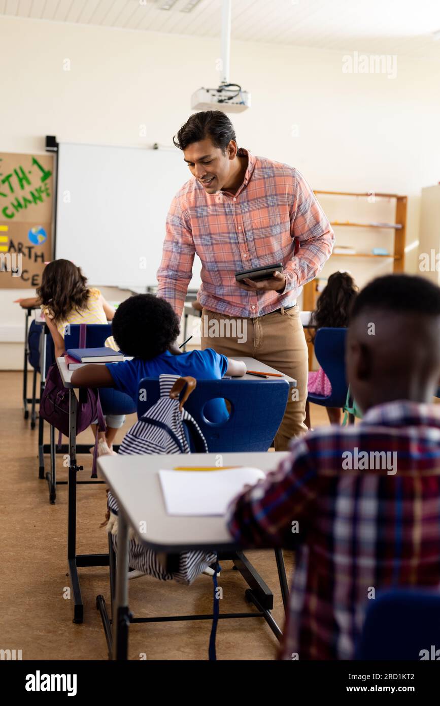 Diverse male teacher with tablet and elementary schoolchildren sitting ...