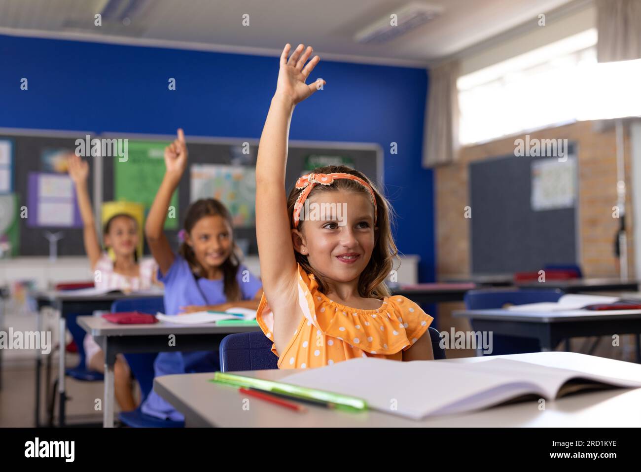 Happy diverse schoolgirls sitting at desks and raising hands in ...