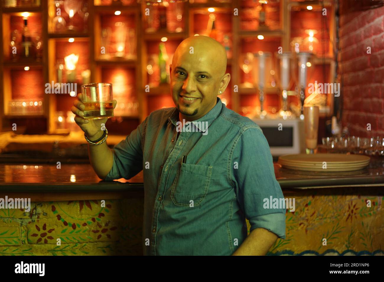 Young and flamboyant handsome bald guy standing on bar counter. Attractive man enjoying in city club interior at night club. Holding whiskey. Stock Photo