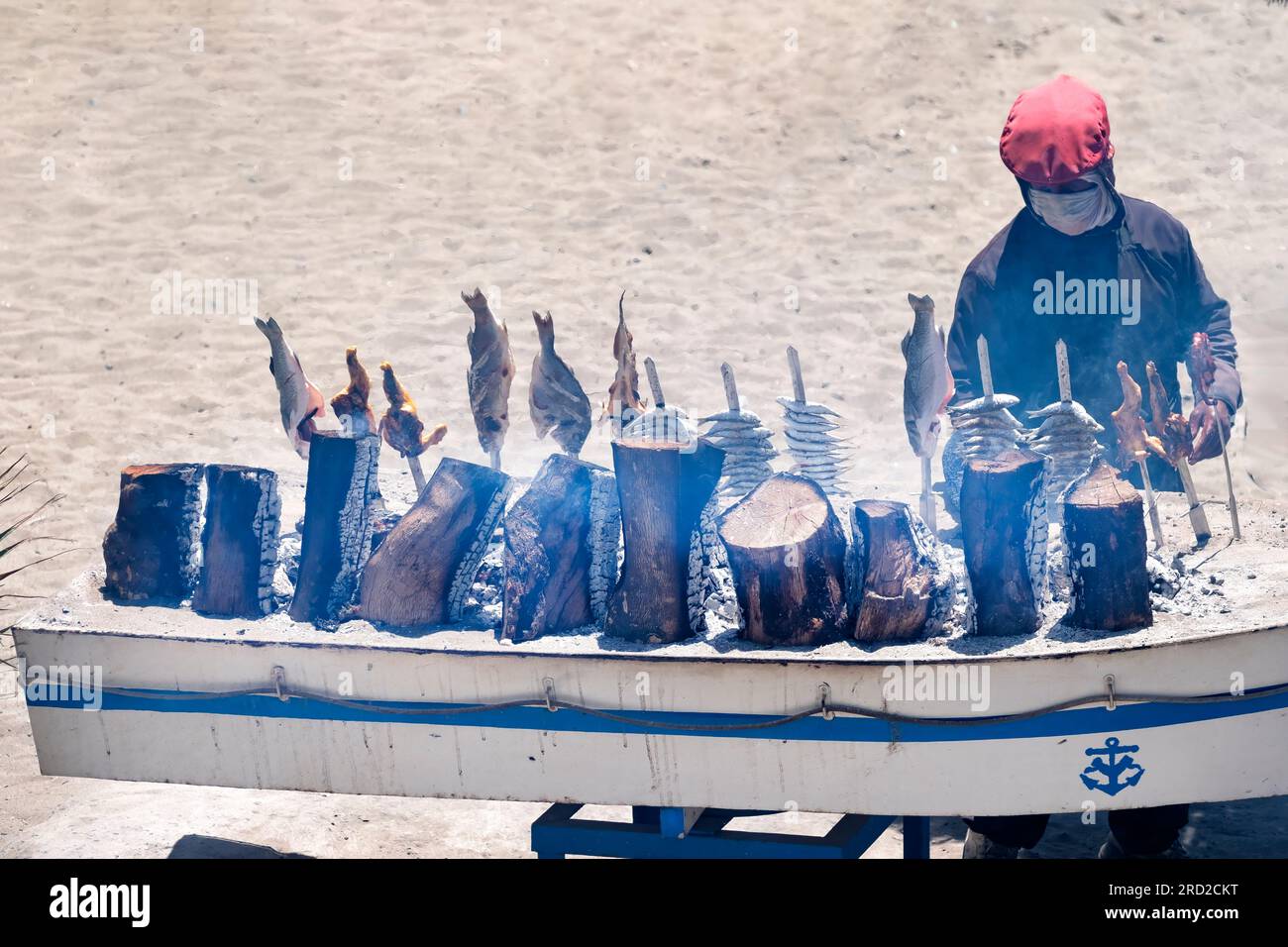A chef cooking fish, sardines and sea bass, on skewers over an open air wood fired barbeque. The BBQ, in a boat, is on the beach on the Costa Del Sol Stock Photo