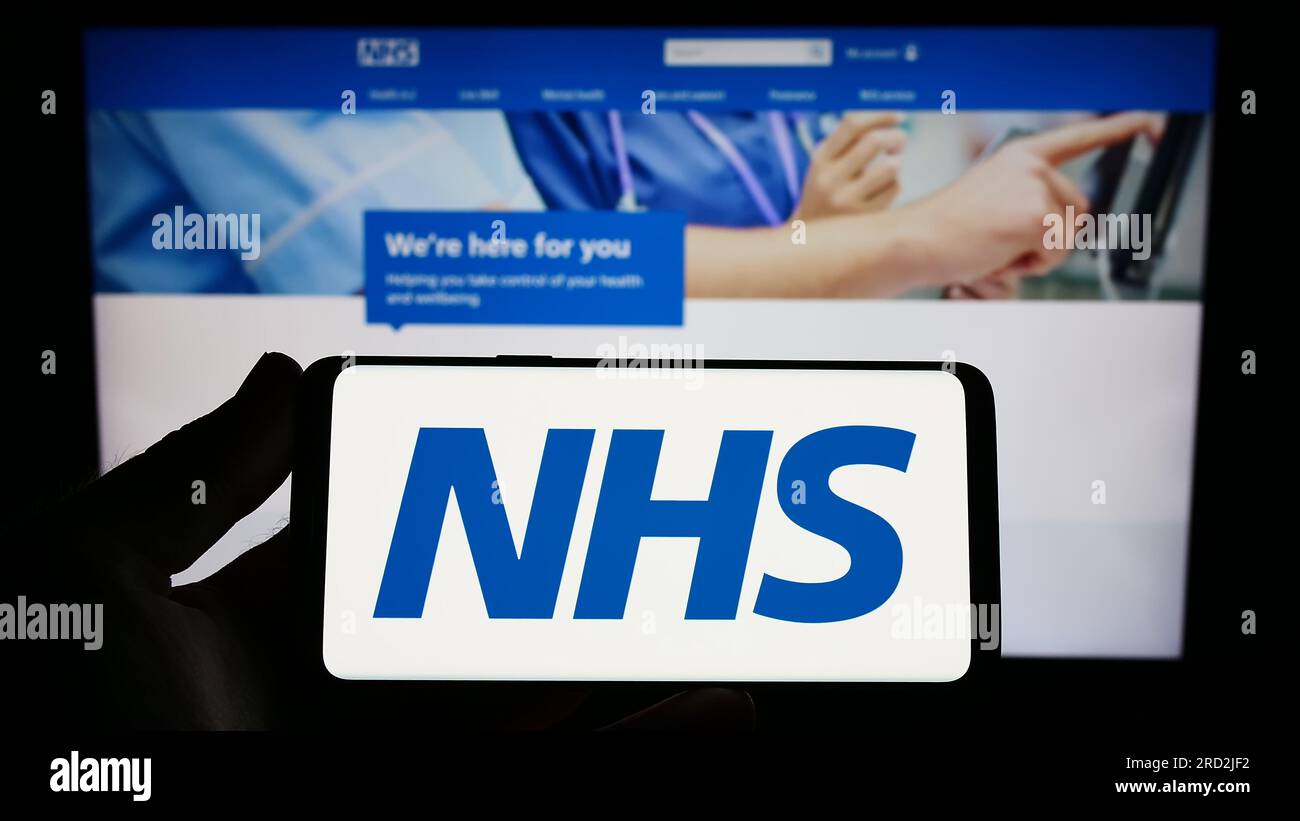 Person holding cellphone with logo of British National Health Service (NHS) on screen in front of webpage. Focus on phone display. Stock Photo