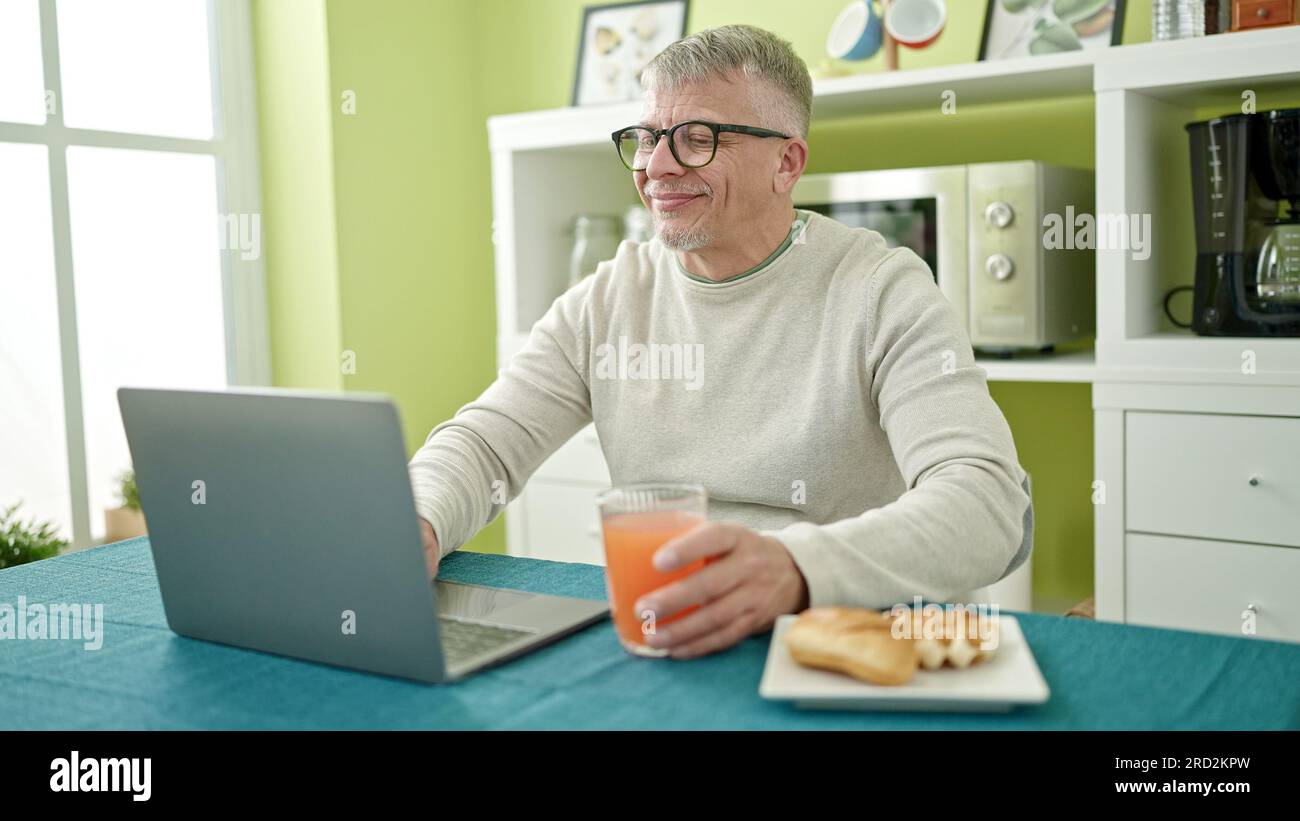 Middle age grey-haired man having breakfast using laptop at home Stock Photo