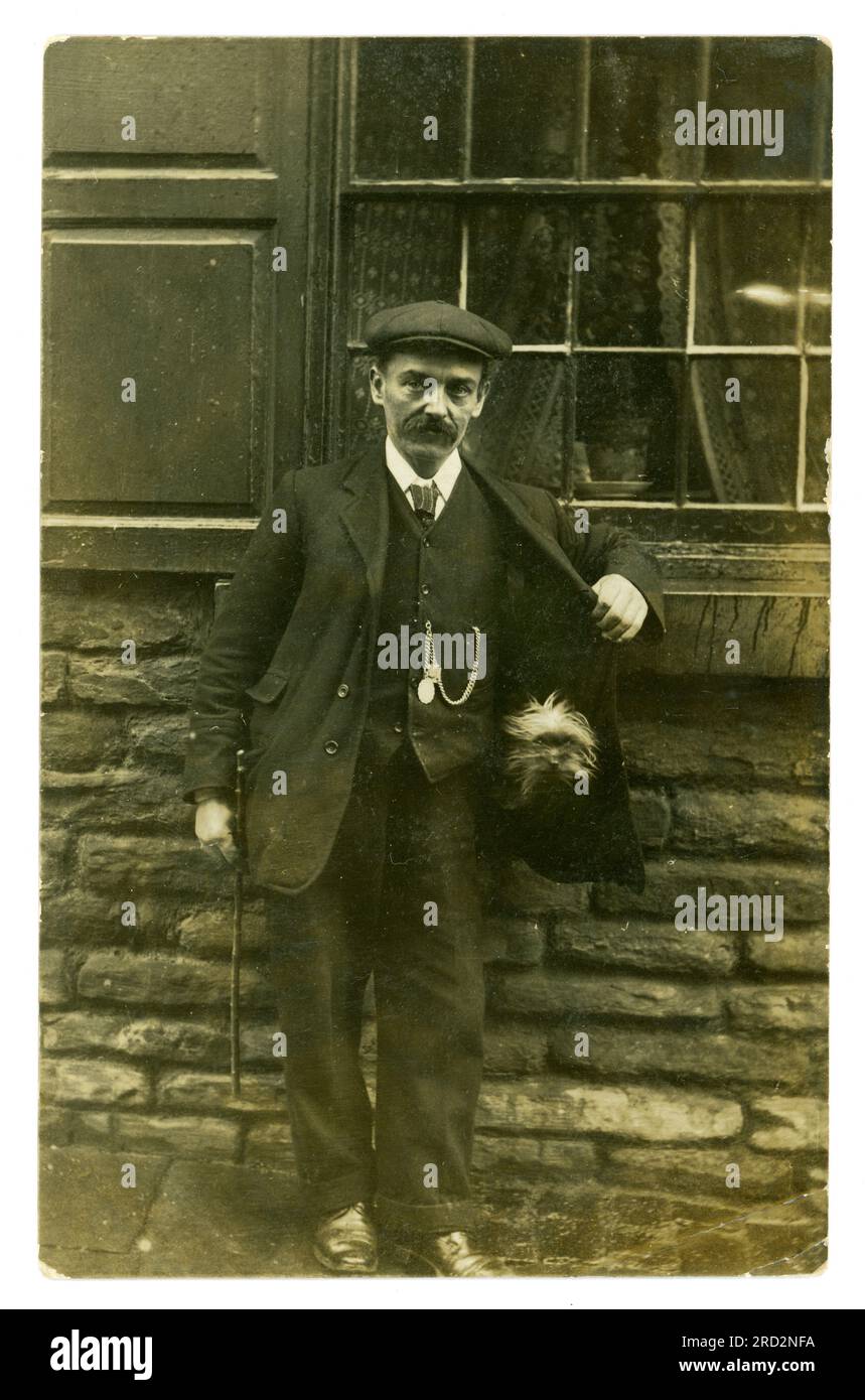 Original, charming early 1920's postcard of working class man with cute toy or small Yorkshire terrier dog or puppy inside coat pocket outside a building possibly a pub. He wears a  Maltese Cross on a watch fob chain, possibly a WW1 veteran or fireman. Possibly Yorkshire, England, U.K. Stock Photo