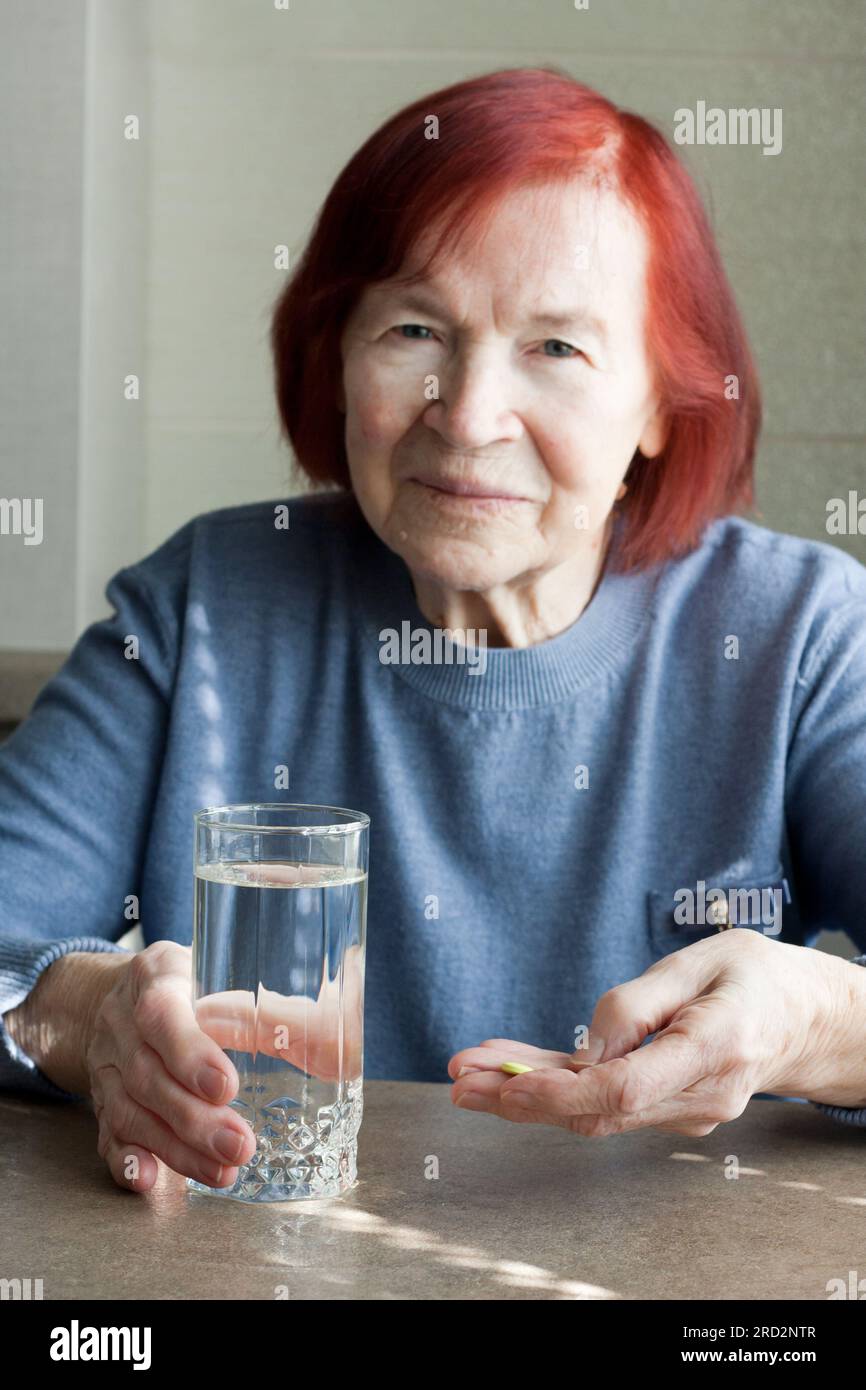 elderly woman holding pills and glass of fresh water, takeing medicines or vitamins. Blurred portrait of sad old woman with red hair sitting at table Stock Photo