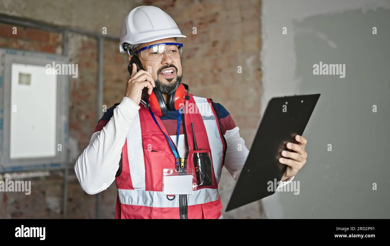 Young latin man builder talking on smartphone reading document at construction site Stock Photo