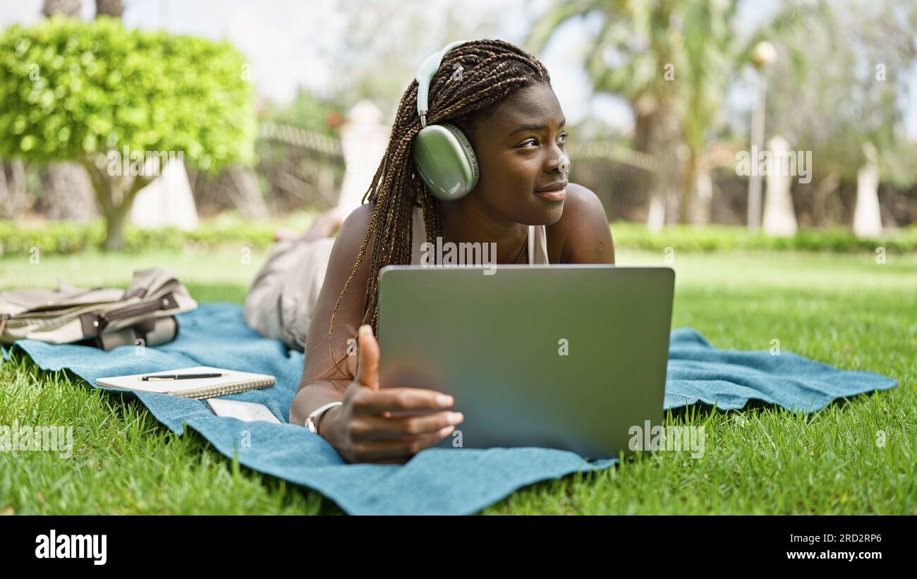 African american woman student using laptop and headphones lying on ...