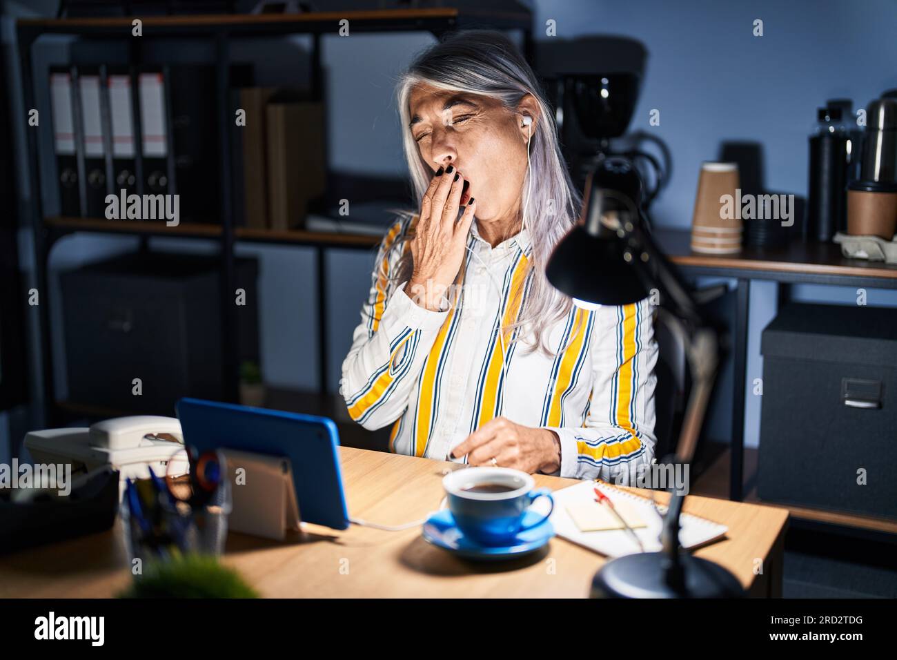 Middle age woman with grey hair working at the office at night bored ...