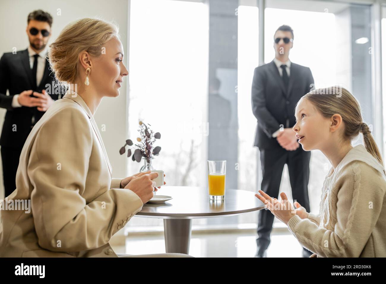 security service, private safety concept, preteen girl talking with blonde mother over drinks in cafe, bodyguards standing on blurred background, pers Stock Photo