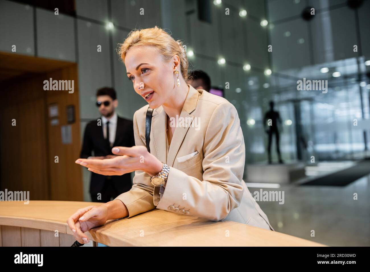 blonde woman in suit gesturing while talking at reception desk, personal security service concept, bodyguards in suits standing on blurred background, Stock Photo