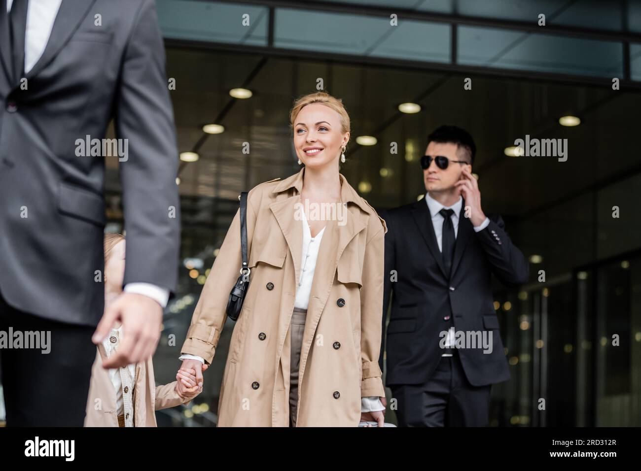professional bodyguards protecting smiling blonde woman and preteen kid, successful mother and daughter in trench coats standing near hotel, safety an Stock Photo