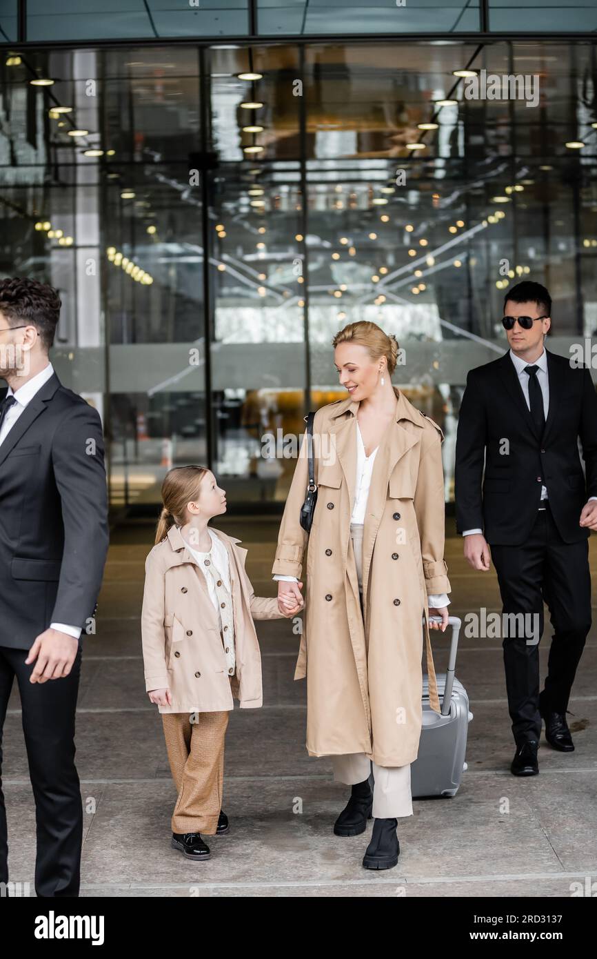 professional bodyguards protecting successful woman and preteen kid, happy blonde mother and daughter in trench coats standing near hotel, safety and Stock Photo