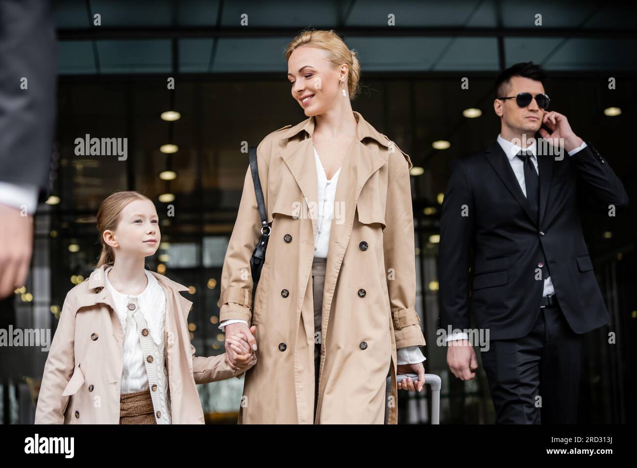 professional bodyguards protecting happy woman and preteen kid, successful blonde mother and daughter in trench coats standing near hotel, safety and Stock Photo