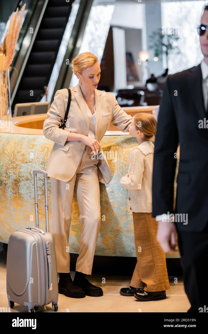 personal security concept, blonde woman standing at reception desk and talking to preteen daughter, rich lifestyle, family travel, bodyguard in suit a Stock Photo