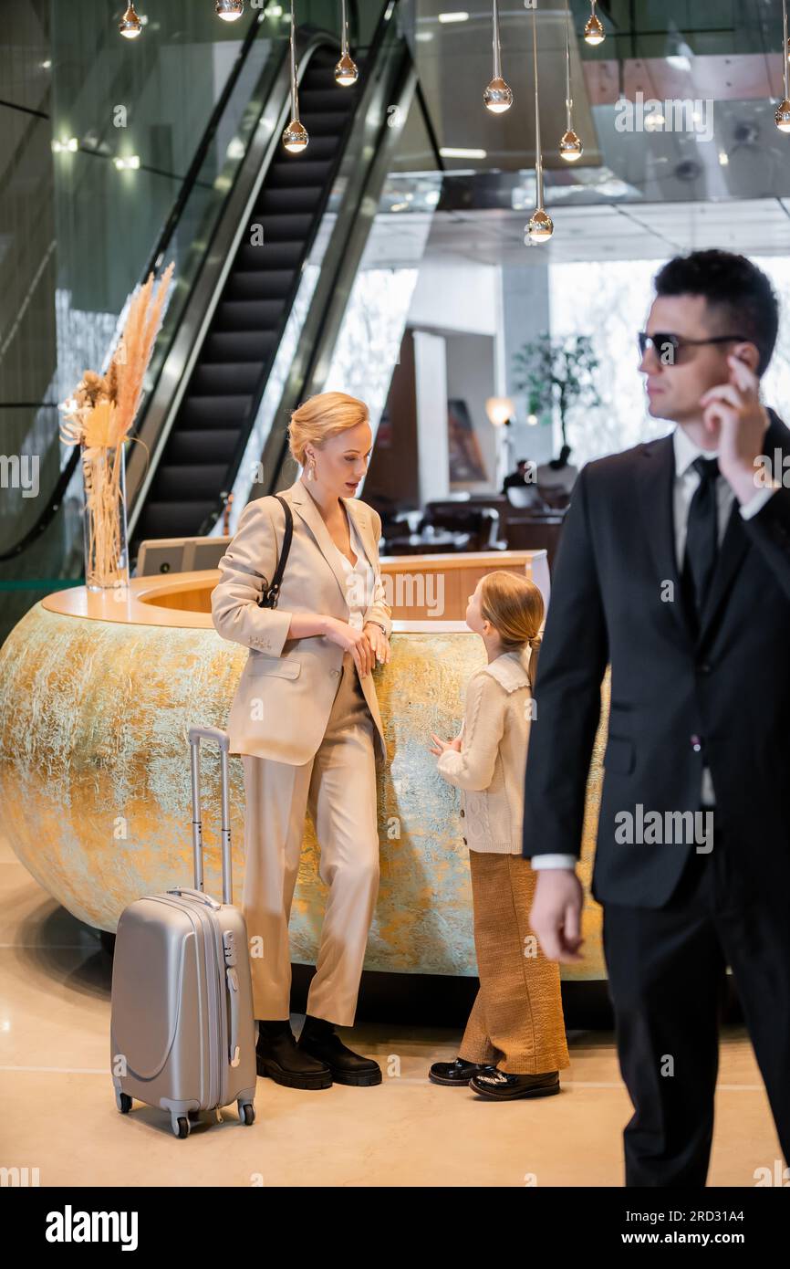 personal safety concept, blonde woman standing at reception desk with preteen daughter, rich lifestyle, family travel, bodyguard in suit and sunglasse Stock Photo