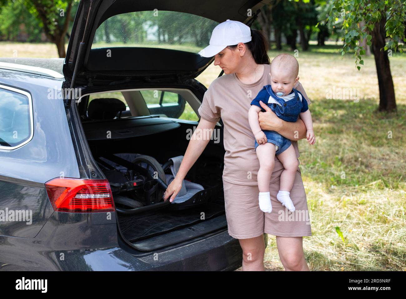 A young mother pushes a pram into the trunk of a car with a baby in her ...
