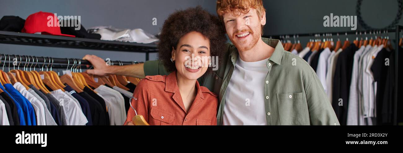 Smiling young multiethnic craftspeople looking at camera while standing together near clothes on hangers in print studio, young small business owners Stock Photo