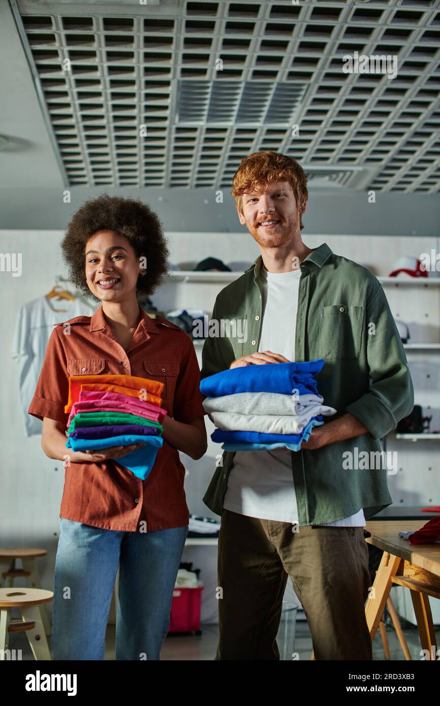 Cheerful young interracial craftspeople holding clothes and looking at camera while standing in blurred print studio at background, ambitious young en Stock Photo
