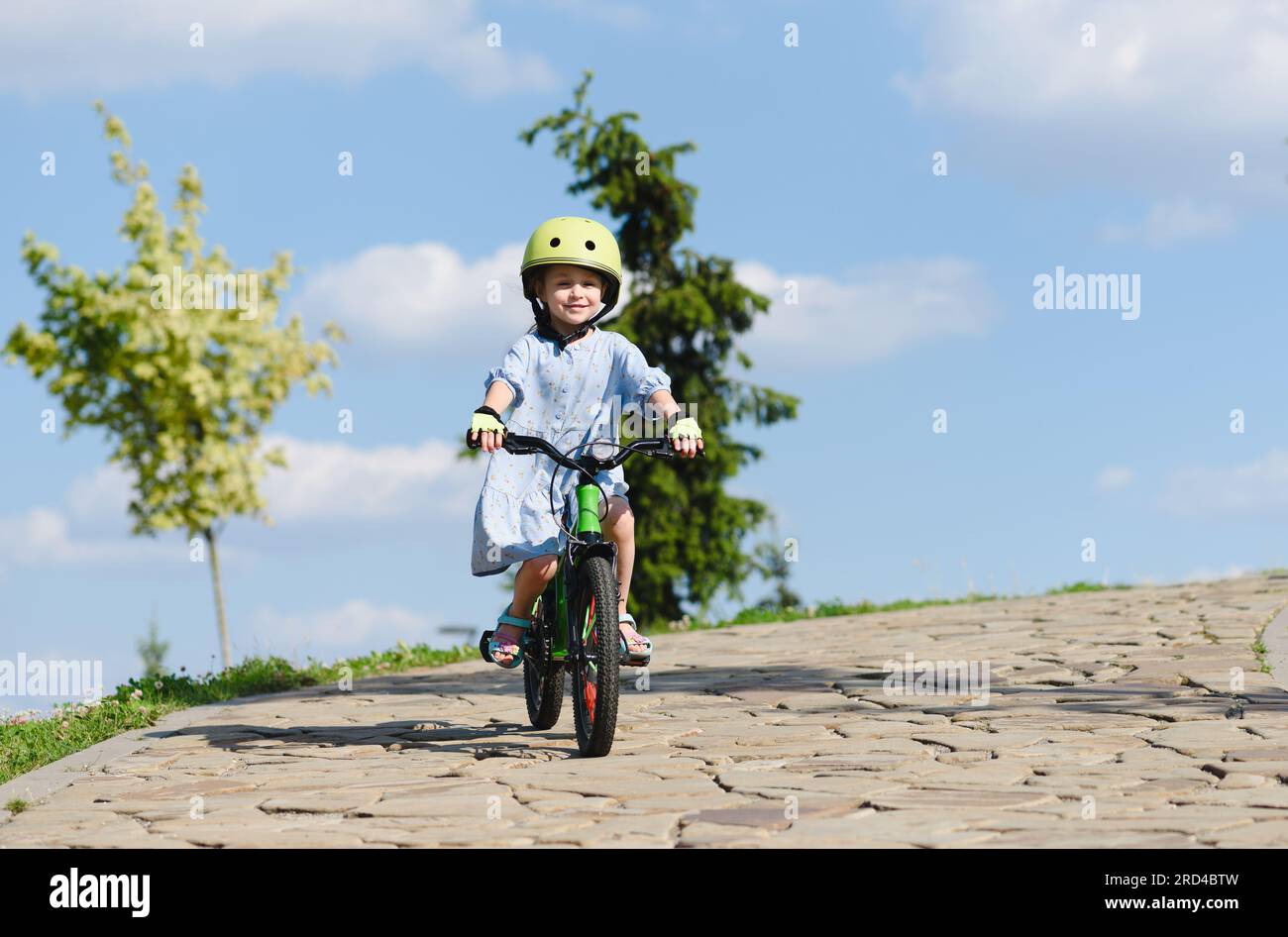 Happy small girl riding bike downhill in park with blue sky in background Stock Photo