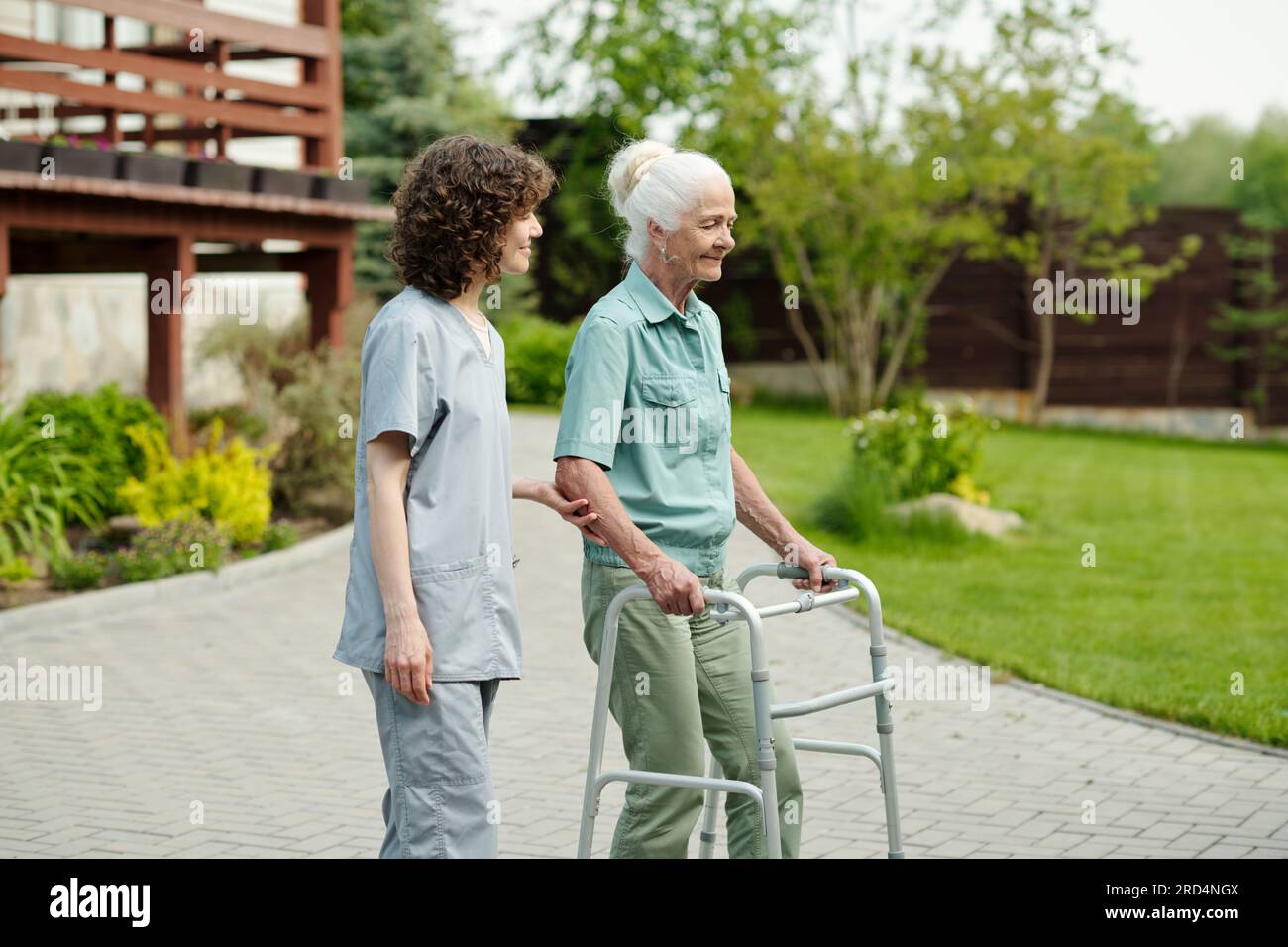 Young caregiver supporting hand of senior patient of retirement home using walker during stroll in the garden with green lawns and trees Stock Photo