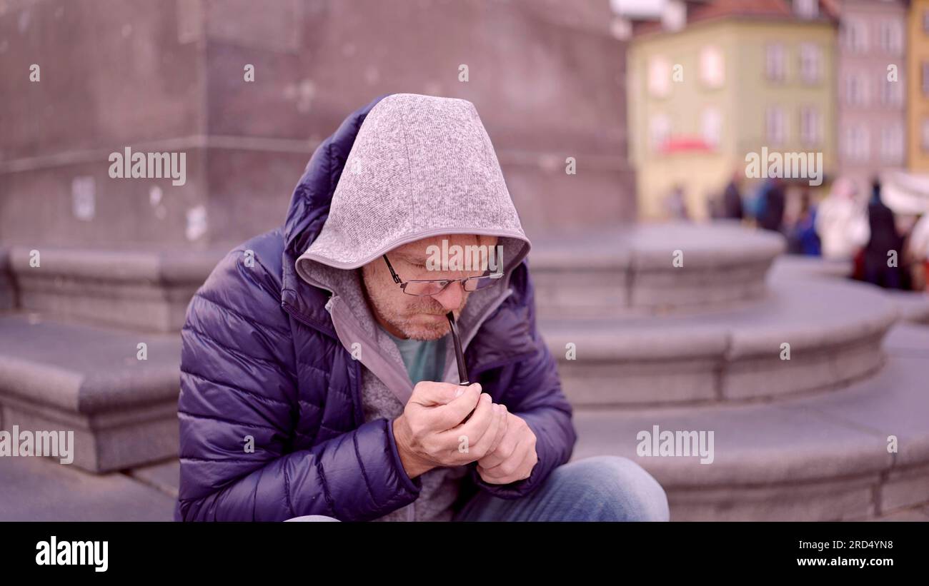 Adult man Ð² Ð¾Ñ‡ÐºÐ°Ñ… sitting on square and lights up at a tobacco pipe in the Palace Square, Warsaw Old Town Stock Photo