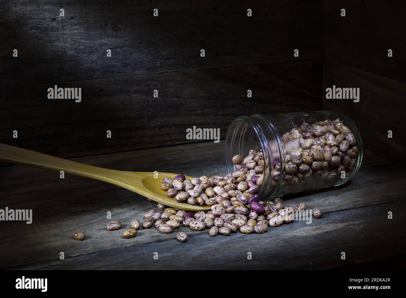 A still life of a pile of raw, dry, uncooked, beige, brown Borlotti beans spilling from a glass jar with wooden spoon in the corner of a wooden box Stock Photo