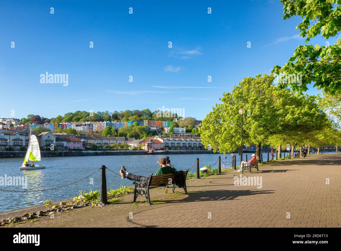 10 May 2023: Bristol, UK - A beautiful evening at Bristol Docks, young couple sitting on a bench, colourful houses,fresh green trees, blue sky, sail b Stock Photo
