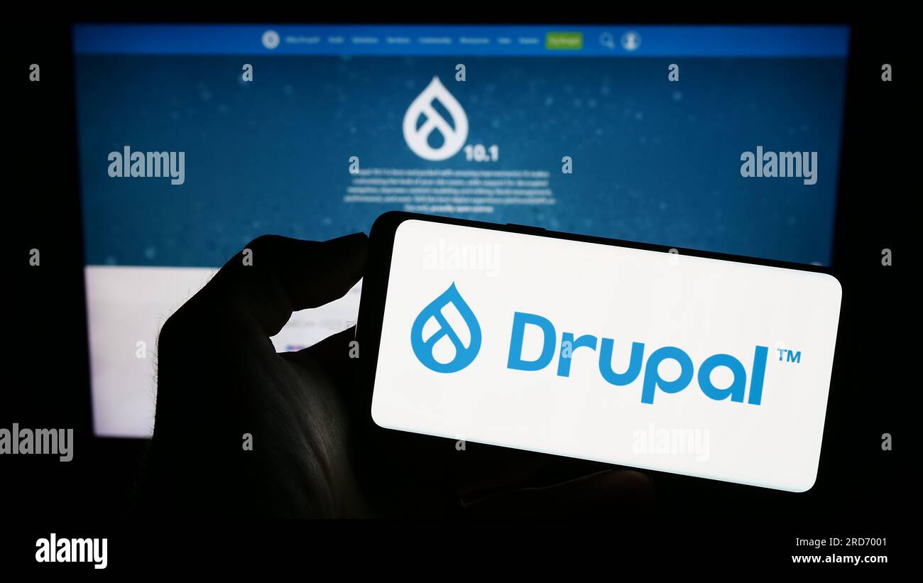 Person holding cellphone with logo of content management system Drupal on screen in front of webpage. Focus on phone display. Stock Photo