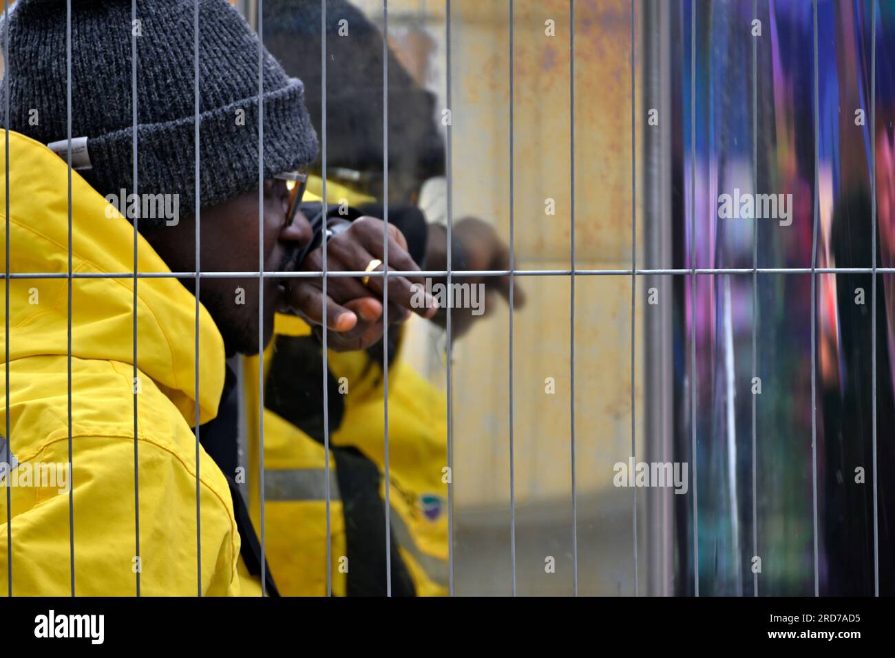 Man in yellow waterproof coat with hat behind crowed control wire fence with reflections behind Stock Photo