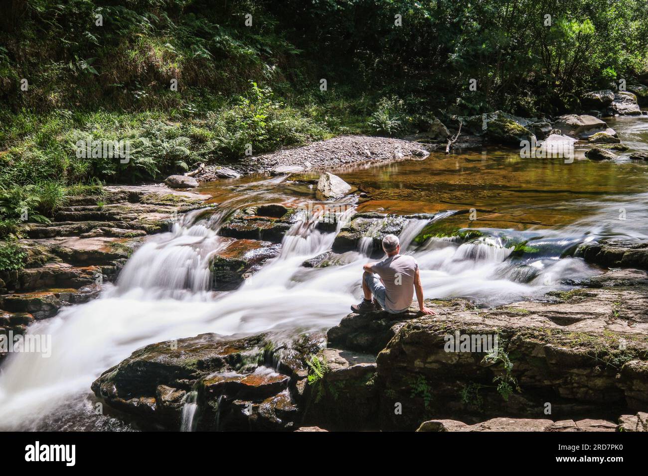 Watersmeet, Lynmouth, Devon, UK. 19th July 2023. UK Weather. A walker talks a rest in the sunshine  by the river at Watersmeet, where the Hoar Oak water joins the East Lyn river. Credit Simon Maycock / Alamy Live News. Stock Photo