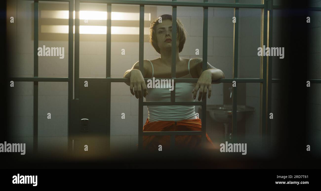 Female prisoner in orange uniform holds hands on bars, stands in jail cell, looks at camera. Woman criminal serves imprisonment term in prison. Detention center or correctional facility. Portrait. Stock Photo