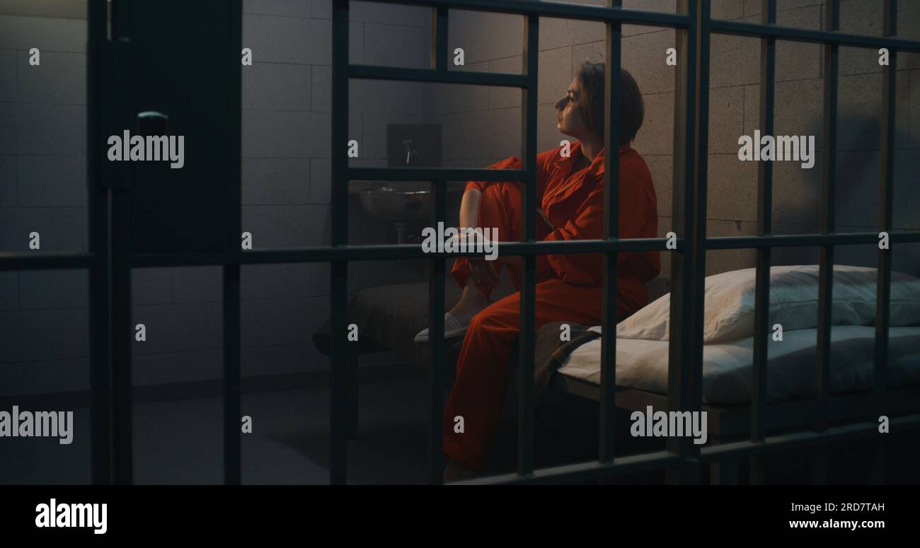 Depressed female prisoner, inmate in orange uniform sits on bed in prison cell. Woman criminal serves imprisonment term for crime in jail. Detention center or correctional facility. Justice system. Stock Photo