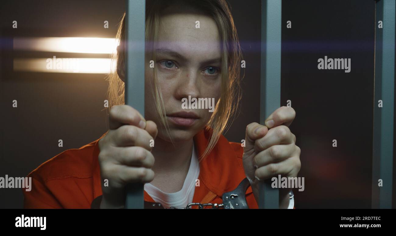 Female prisoner in orange uniform stands in prison cell. Scared woman criminal shakes, holds metal bars by hands. Depressed inmate serves imprisonment term in jail or correctional facility. Portrait. Stock Photo