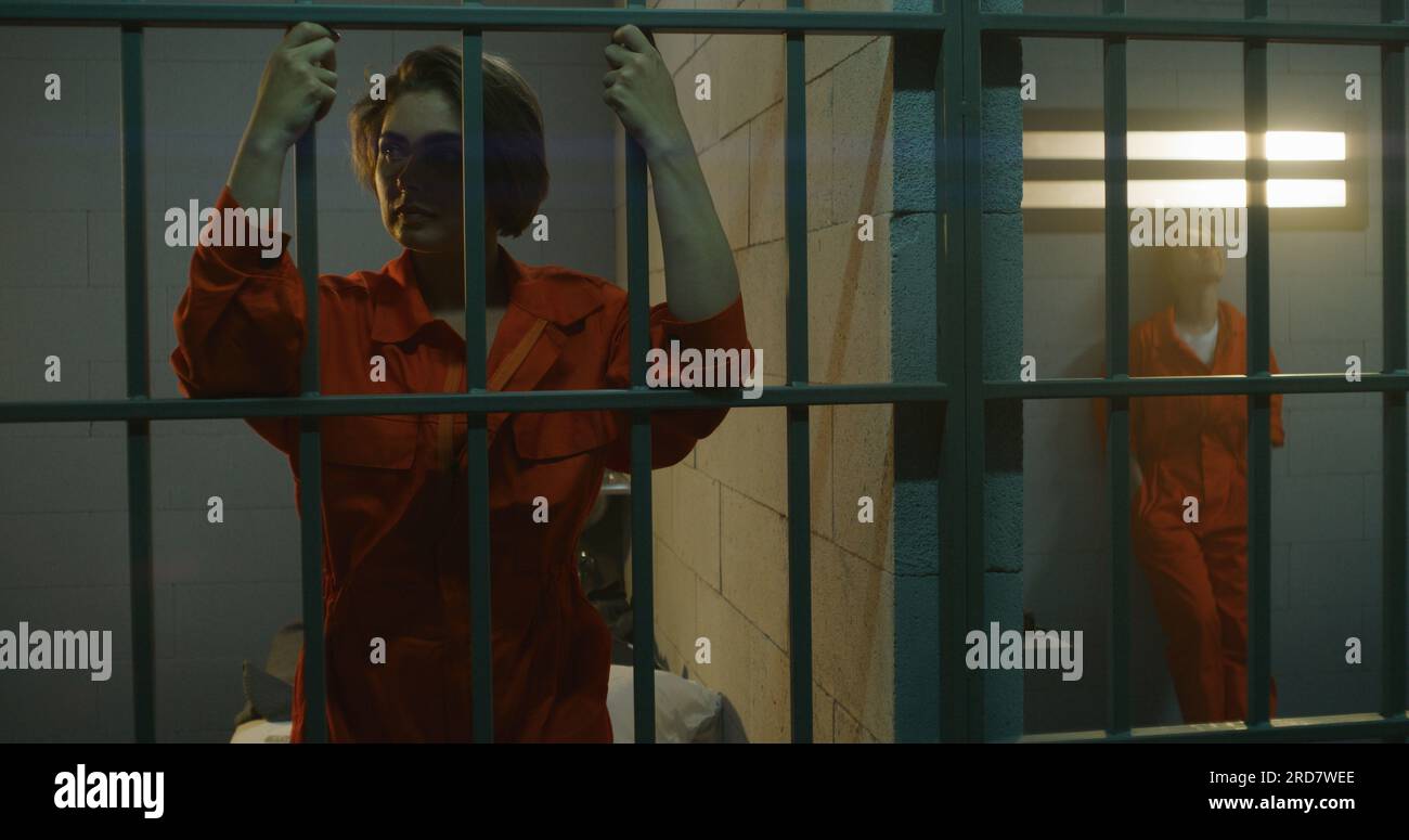 One female prisoner in orange uniform stands behind metal bars, another sits on the bed in