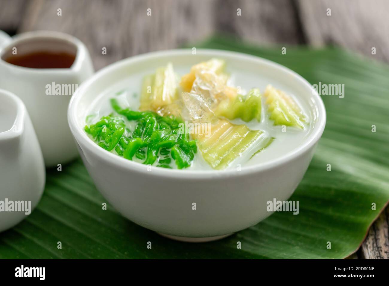 Close up of Lod Chong Thai dessert topped with coconut milk and pandan leaf followed by green banana leaves on the wood background. Stock Photo
