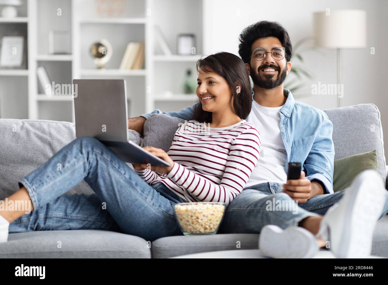 Relaxed eastern couple resting at home, watching tv, using laptop Stock Photo