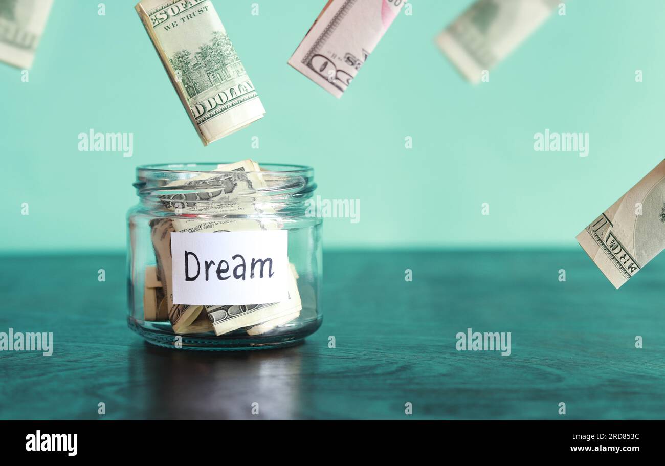 Save money for a dream. Glass jar with dollars on a wooden table. Piggy bank with banknotes and dollars flying around. Copy space. The concept of fina Stock Photo
