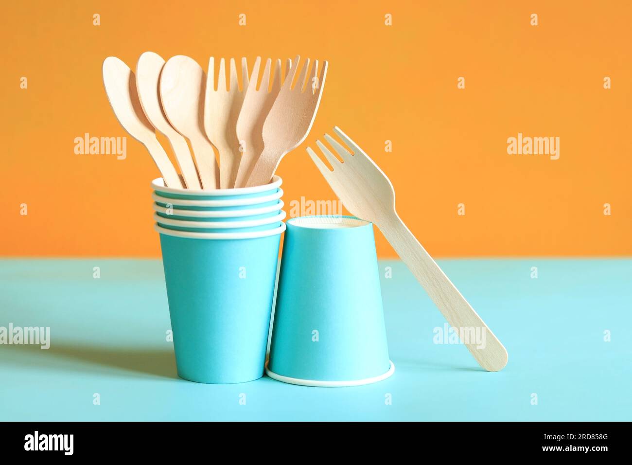 Wooden disposable forks and spoons in a blue paper cup, side view. Eco friendly disposable kitchen utensils on orange background, copy space. Ecologic Stock Photo