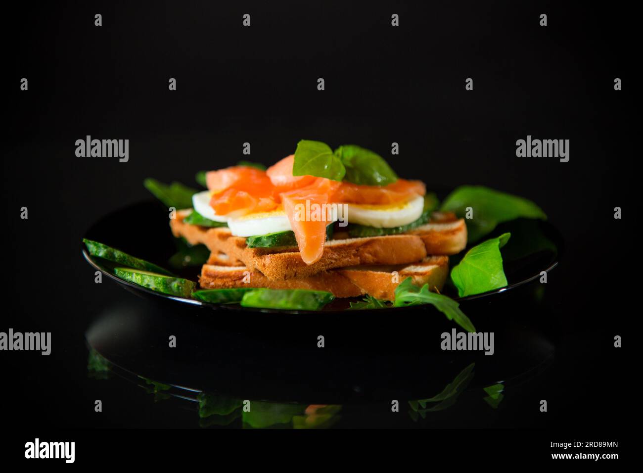 Cooked sandwich with lightly salted red fish, cucumbers and boiled eggs with herbs, isolated on a black background. Stock Photo