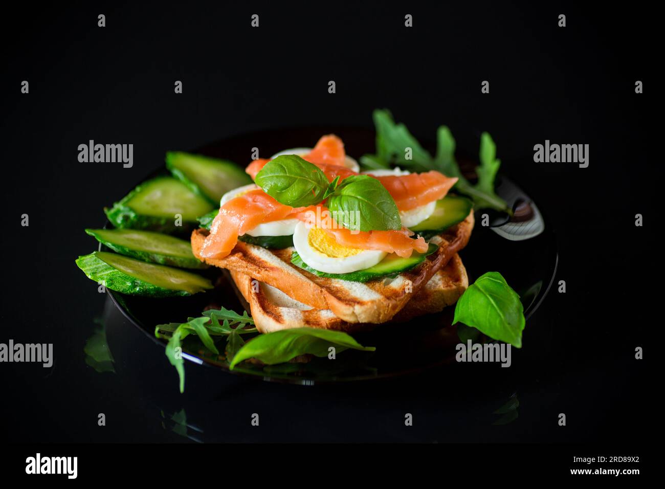Cooked sandwich with lightly salted red fish, cucumbers and boiled eggs with herbs, isolated on a black background. Stock Photo