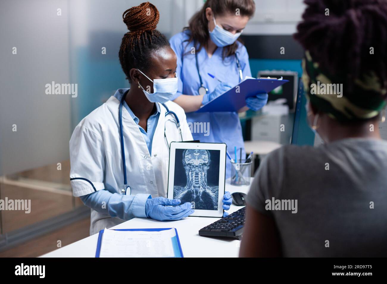 Cervical radiology specialist showing spinal ct scan results on tablet to patient with neck pain. Doctor and nurse with protective workwear gloves and face mask holding x-ray scan at medical checkup Stock Photo