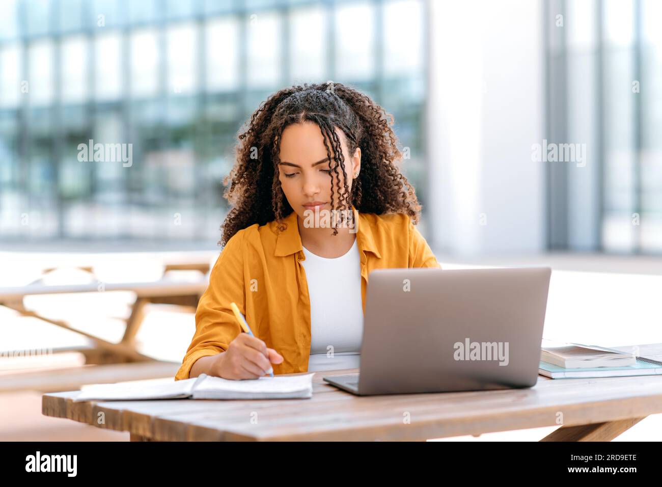 Focused trendy hispanic or brazilian curly haired girl, student or freelancer, sitting outdoors with a laptop, working or studying online, concentrated writing down information in her notepad Stock Photo
