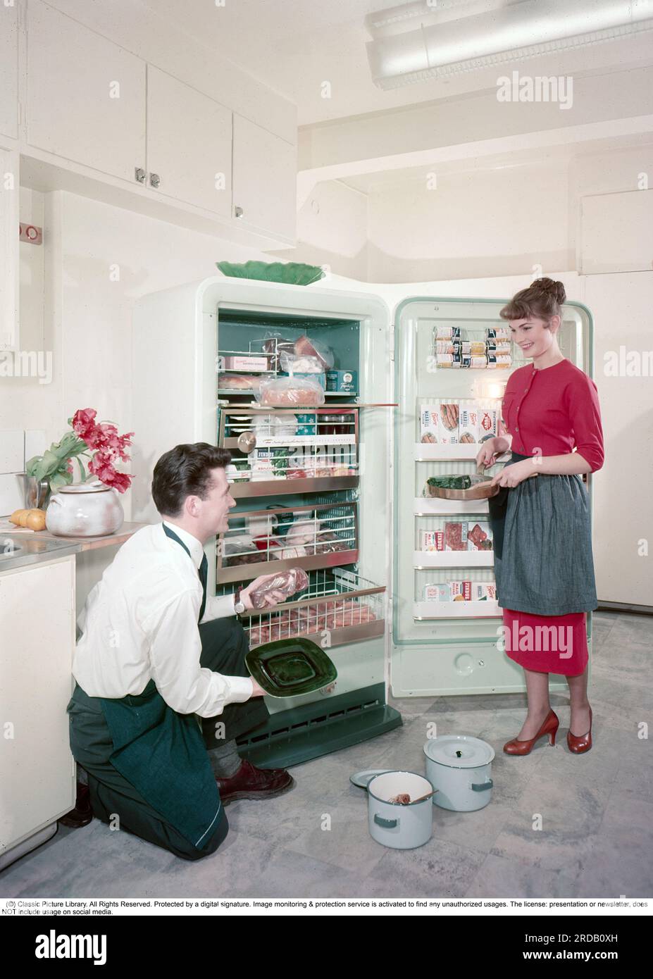 Kitchen from the 1950s. The young Swedish couple in the period kitchen. The door is open to their new freezer where the shelves are filled with frozen goods. Deep-frozen food would be the start of a revolution in household habits and food storage. Many were sceptical, and especially canned food manufacturers considered frozen food to be a passing trend. Sweden 1959. ref BV111-3 Stock Photo