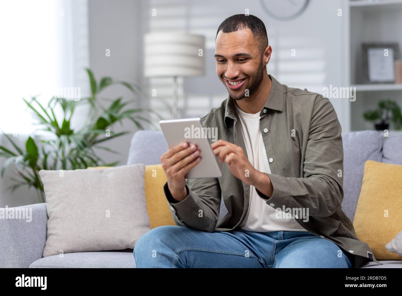 Hispanic smiling young man using tablet at home on sofa. Stock Photo
