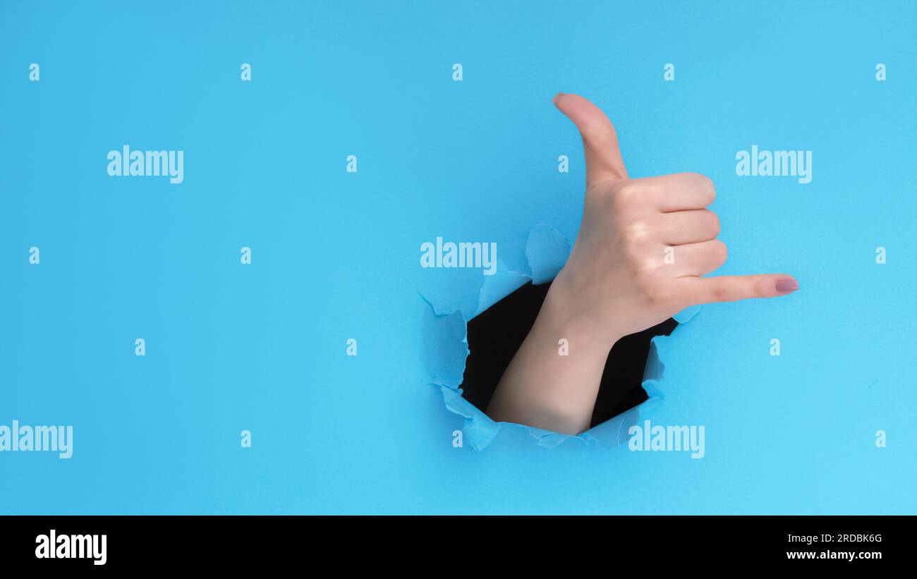 Call gesture. Shaka sign. Hang loose. Female hand showing greeting signal inside breakthrough hole on blue torn paper wall background with empty space Stock Photo