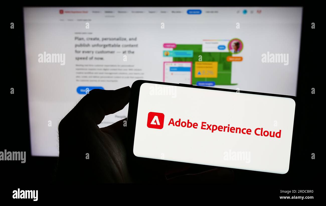 Person holding cellphone with logo of software product Adobe Experience Cloud (AEC) on screen in front of webpage. Focus on phone display. Stock Photo
